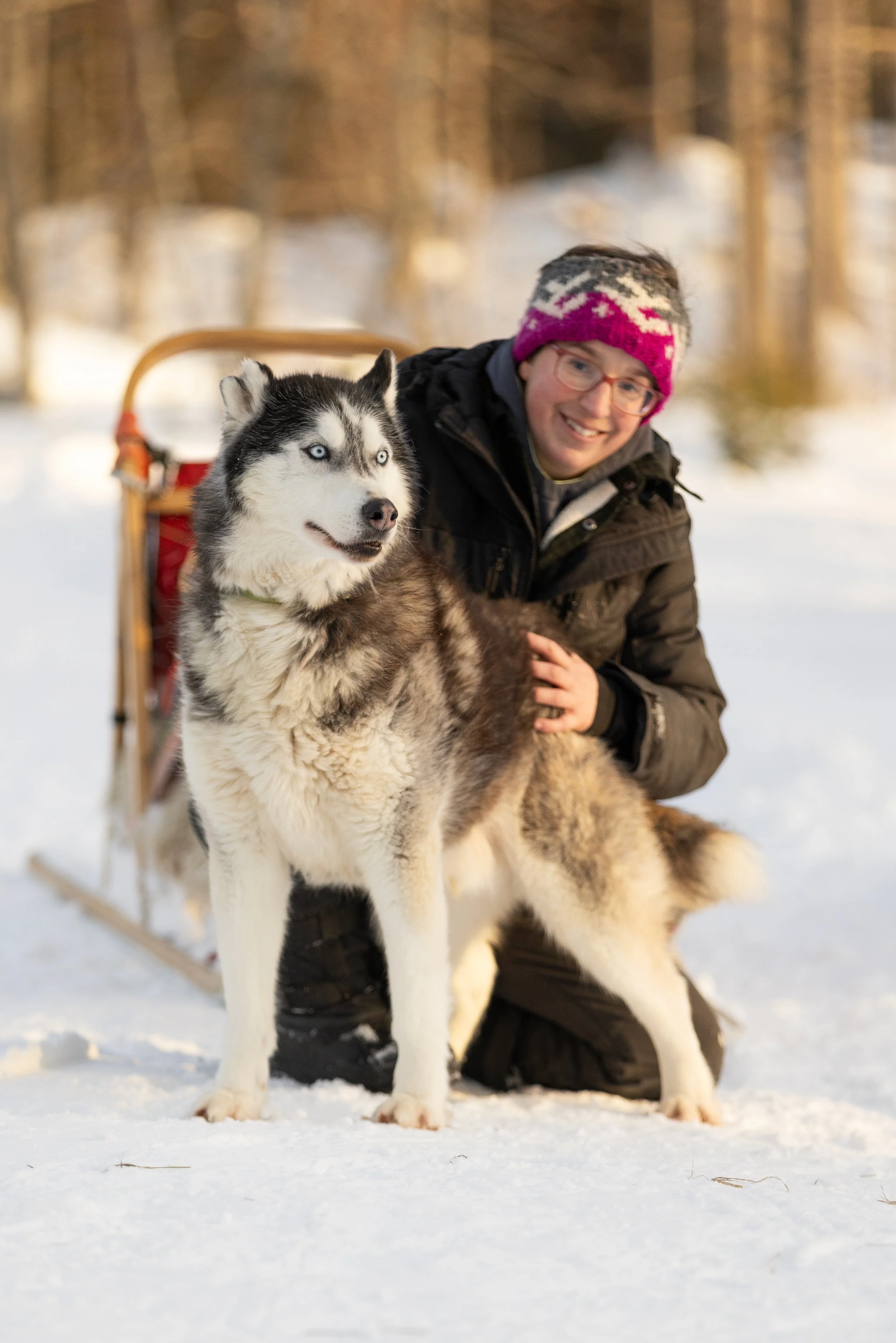 Eine Frau in winterlicher Kleidung neben einem Siberian Husky, im Schnee bei Sonnenuntergang.