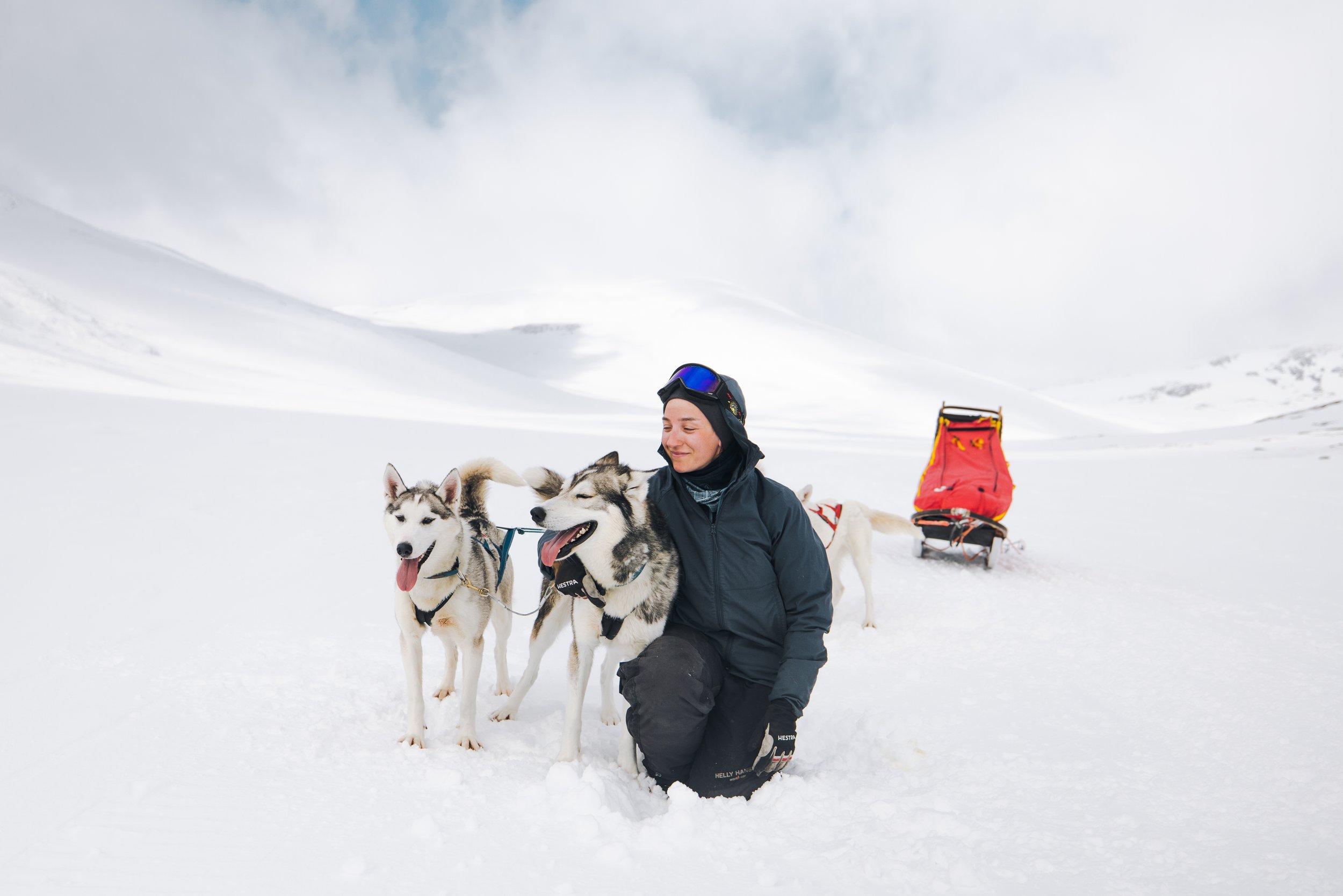 Junge Frau in warmen Kleidung umarmt zwei Siberian Huskies auf verschneiter Landschaft, im Hintergrund Schneelandschaft und Wolken.