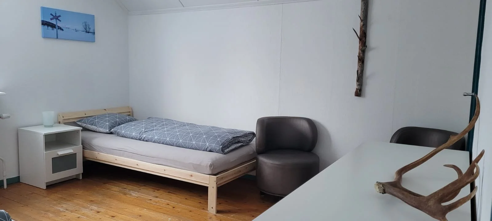 Ein Schlafzimmer mit einem Einzelbett, einem Nachttisch, einem Sessel, einem Tisch und einem Wandbild, das eine verschneite Landschaft zeigt.