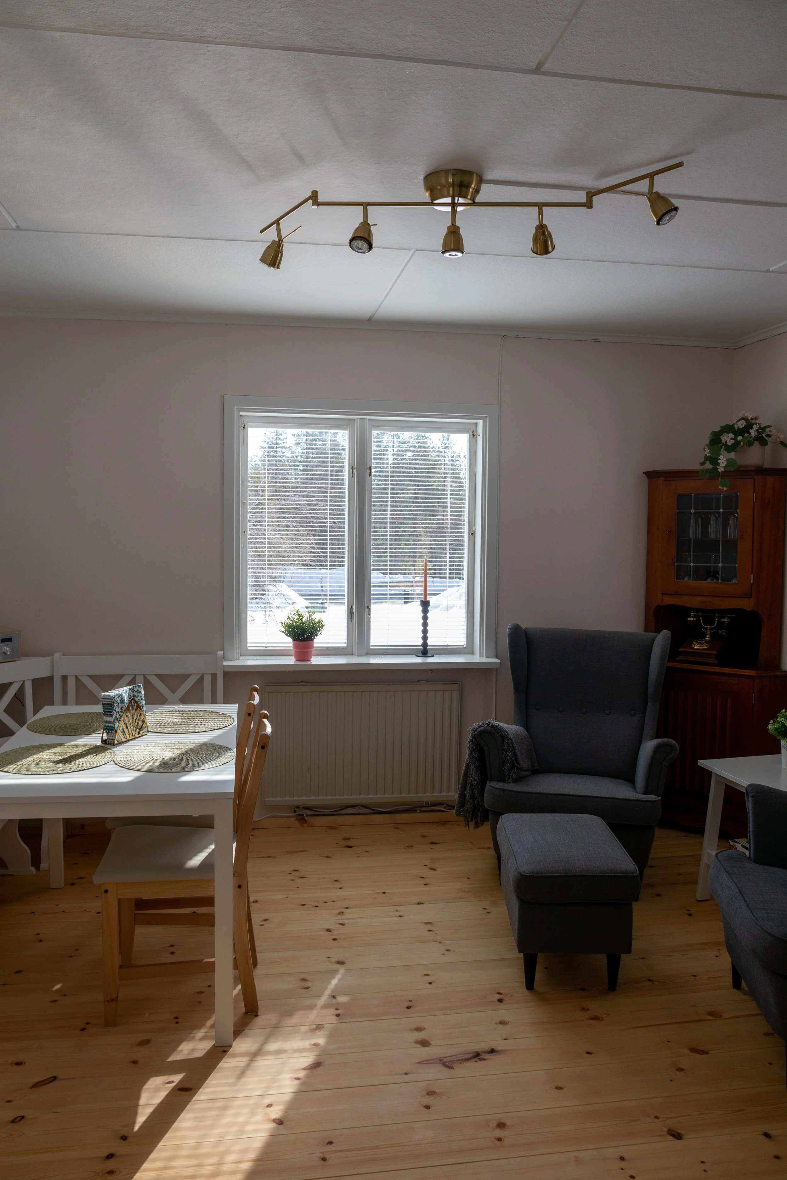 Ein Wohnzimmer mit einem großen Fenster, einem modernen Deckenleuchter, einem weißen Esstisch mit Stühlen, einer grauen Armlehne mit Hocker, einem dunklen Bücherregal und einigen Pflanzen