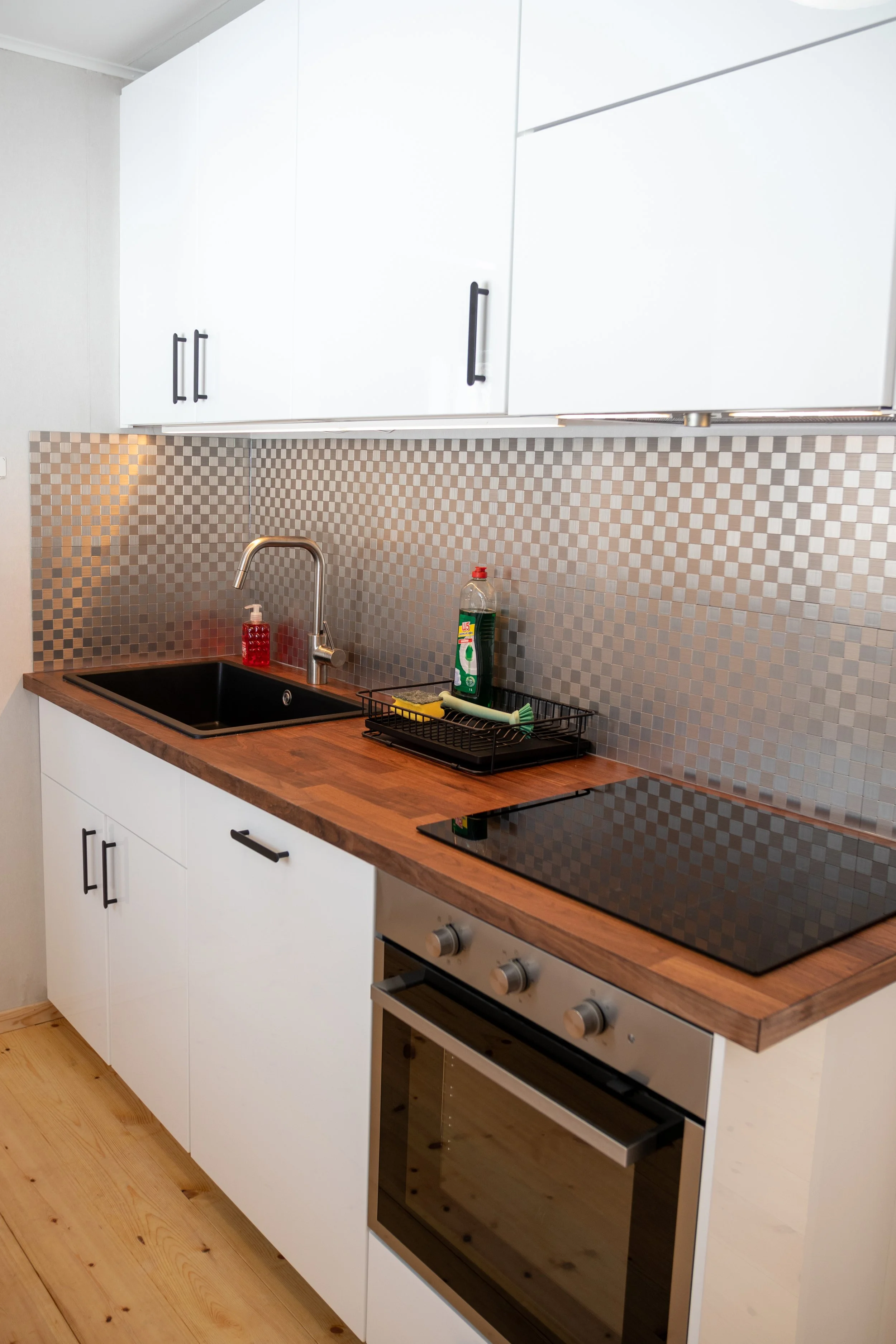 Moderne Küche mit weißen Schränken, Holzarbeitsplatte, schwarzem Spülbecken, Silber-Hahn, Kochfeld und Backofen, wandhängende weiße Schränke, Mosaik-Fliesenwand in beige- und goldfarbenen Tönen, Küchenutensilien und Reinigungsprodukte auf der Arbeitsfläche.