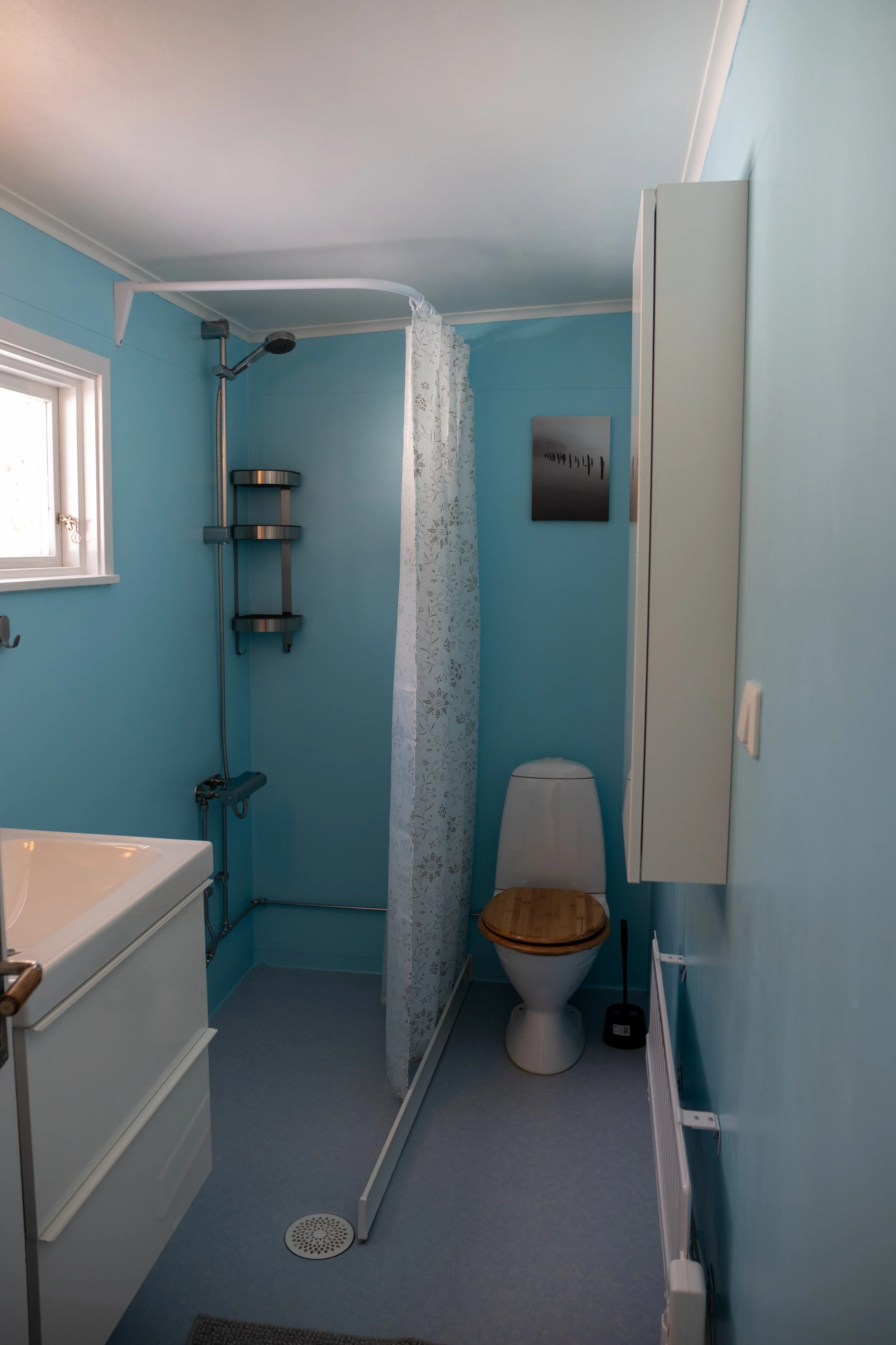 Ein Badezimmer mit hellblauen Wänden, einem Handtuch auf einem Regal, einer Toilette mit Holzsitz, einem weißen Toilettenpapierhalter, einer weißen Spiegelschrank und einer kleinen Fenster. Es gibt auch eine Dusche mit Duschkopf und Duschgelregal.