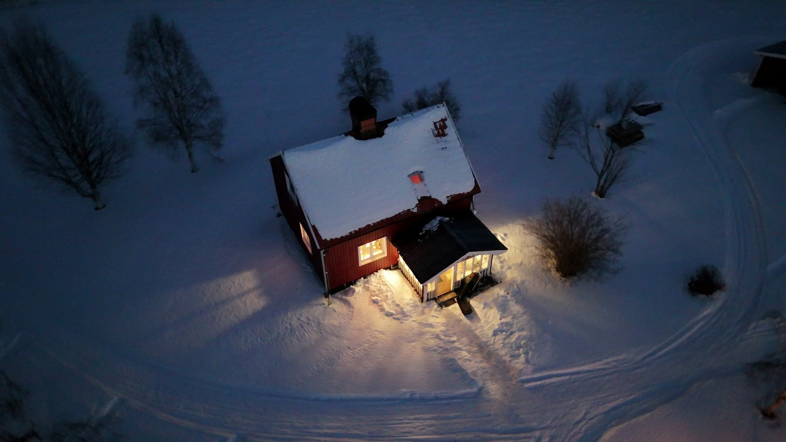 Ein rotes Haus mit Schneedecke auf dem Dach und eingeschalteten Lichtern, umgeben von verschneiten Bäumen in einer Winterlandschaft bei Nacht.