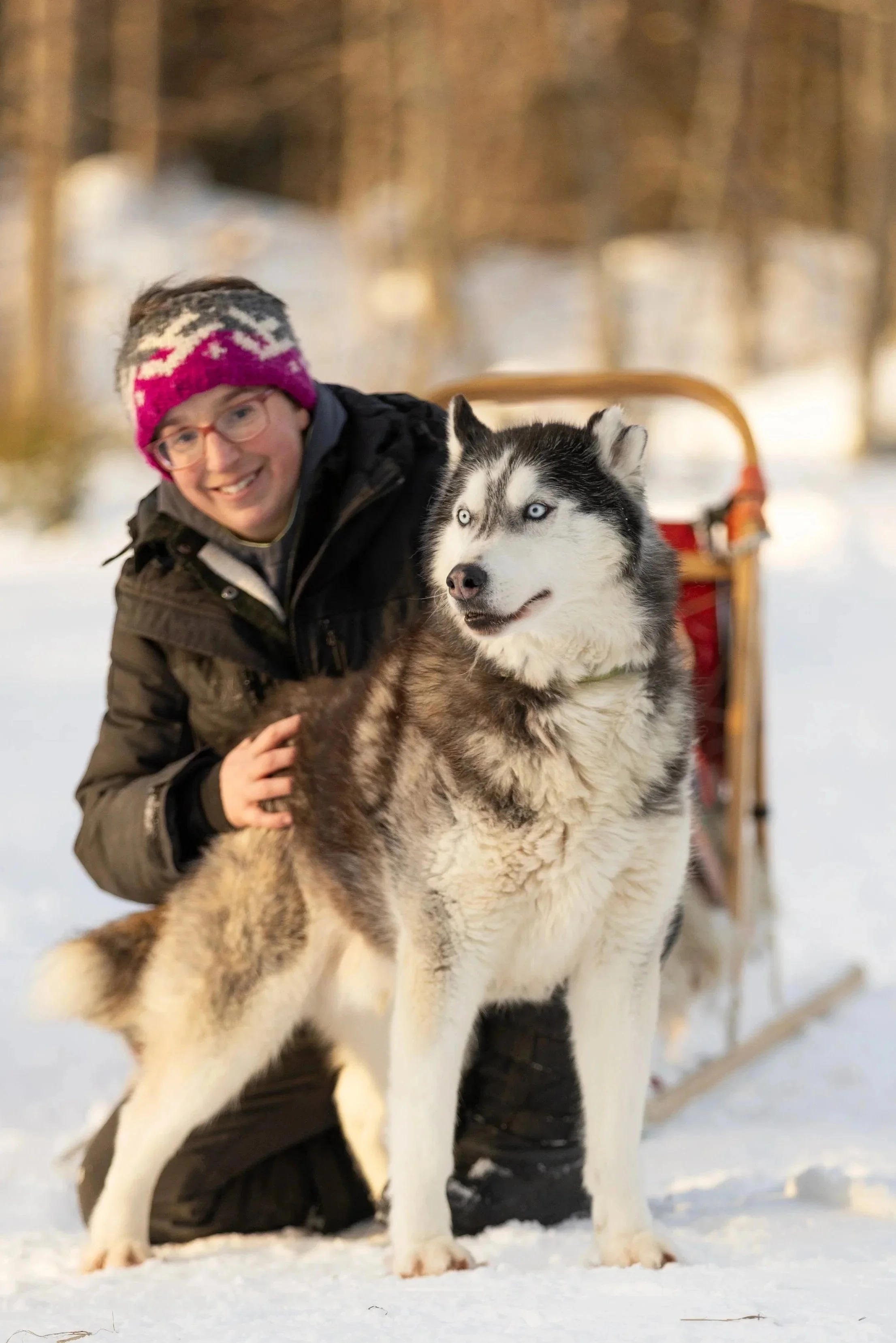 Ein Mann trägt eine Winterjacke, eine bunte Mütze und Brille, und ist neben einem husky Hund im Schnee in einer winterlichen Waldumgebung.