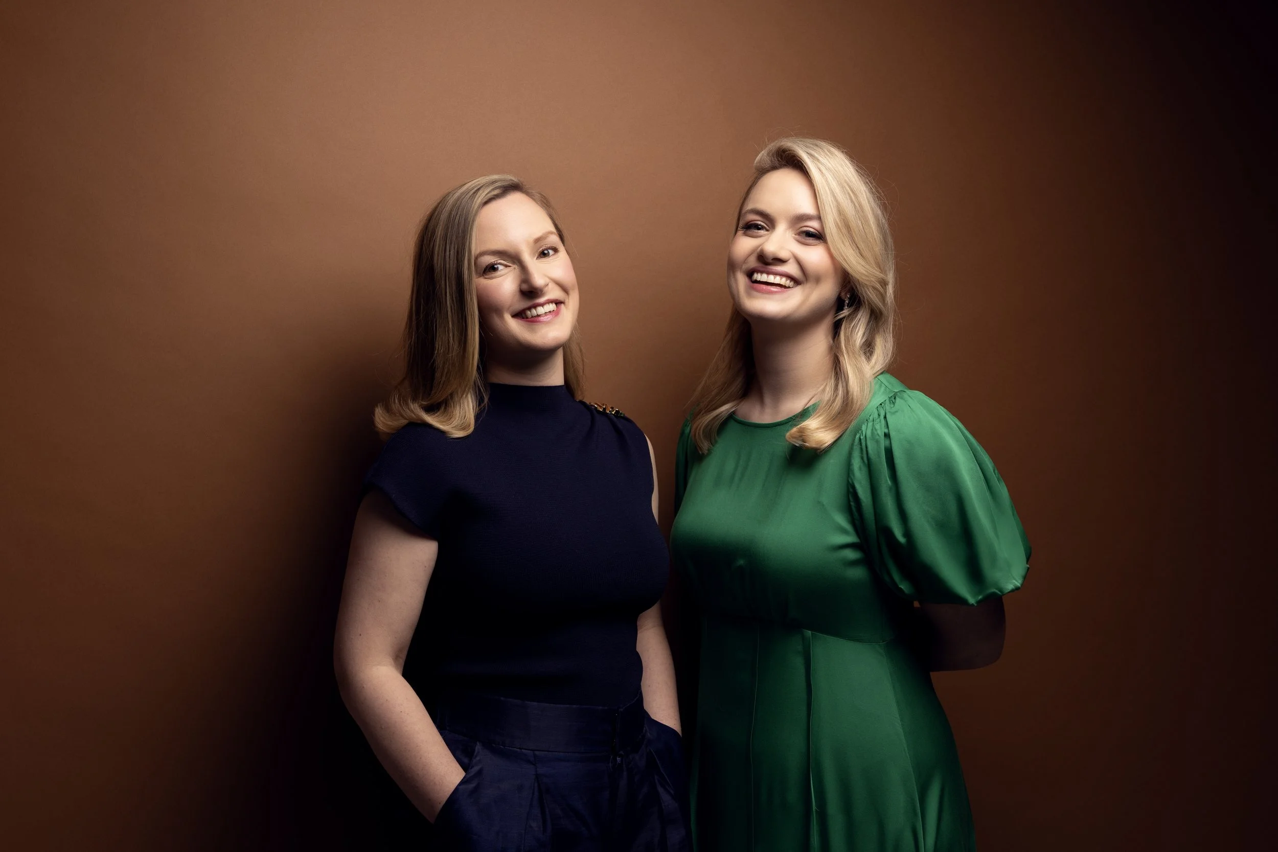 Two women (Natalie Burch on the left, Camilla Harris on the right) smiling in front of a brown background, one wearing a black top and the other in a green dress.