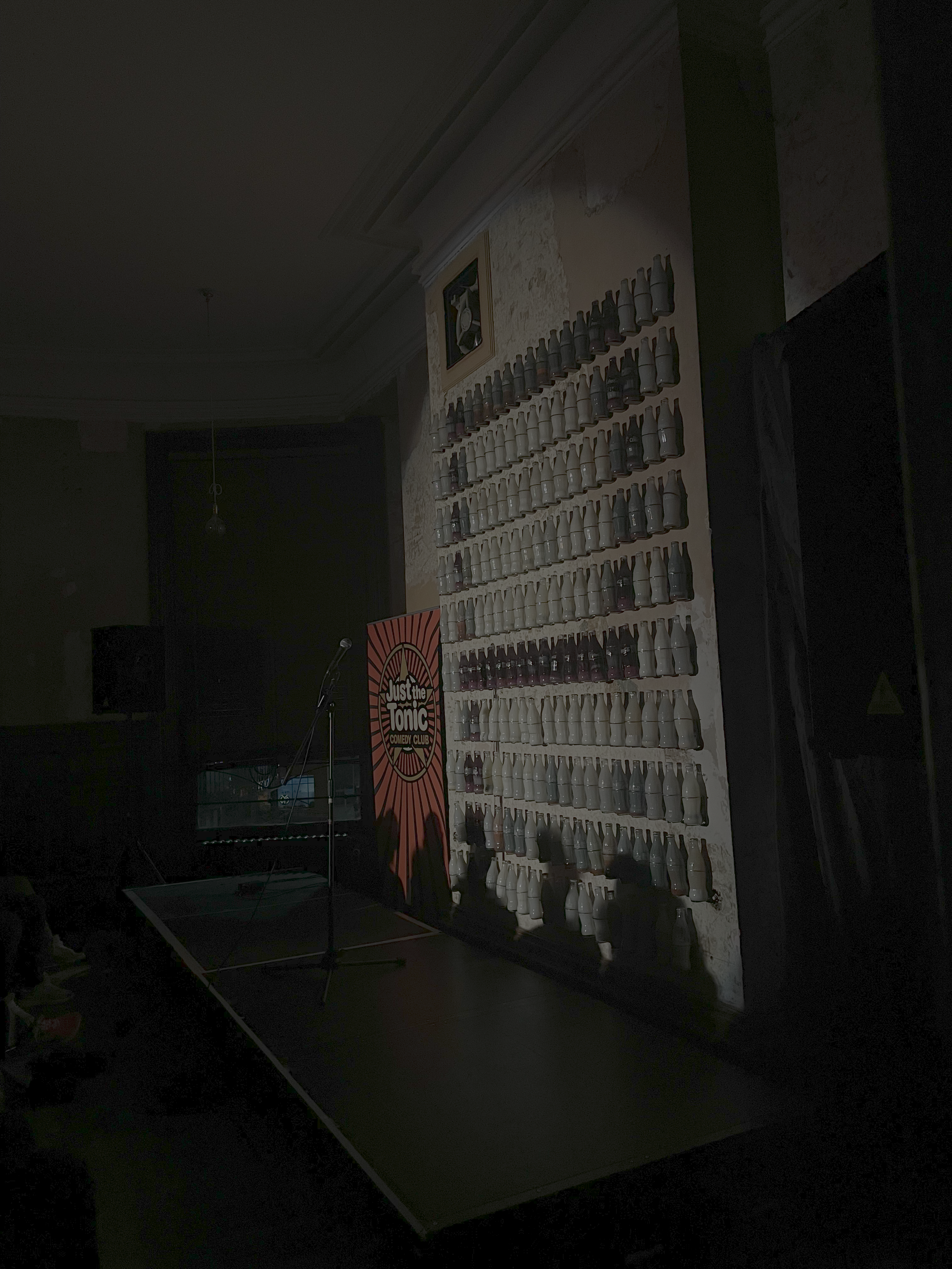 A dimly lit indoor stage features a wall of bowling pins arranged in multiple rows and columns, with a microphone stand in front and a sign for 'Just the Tonic Comedy Club' nearby.