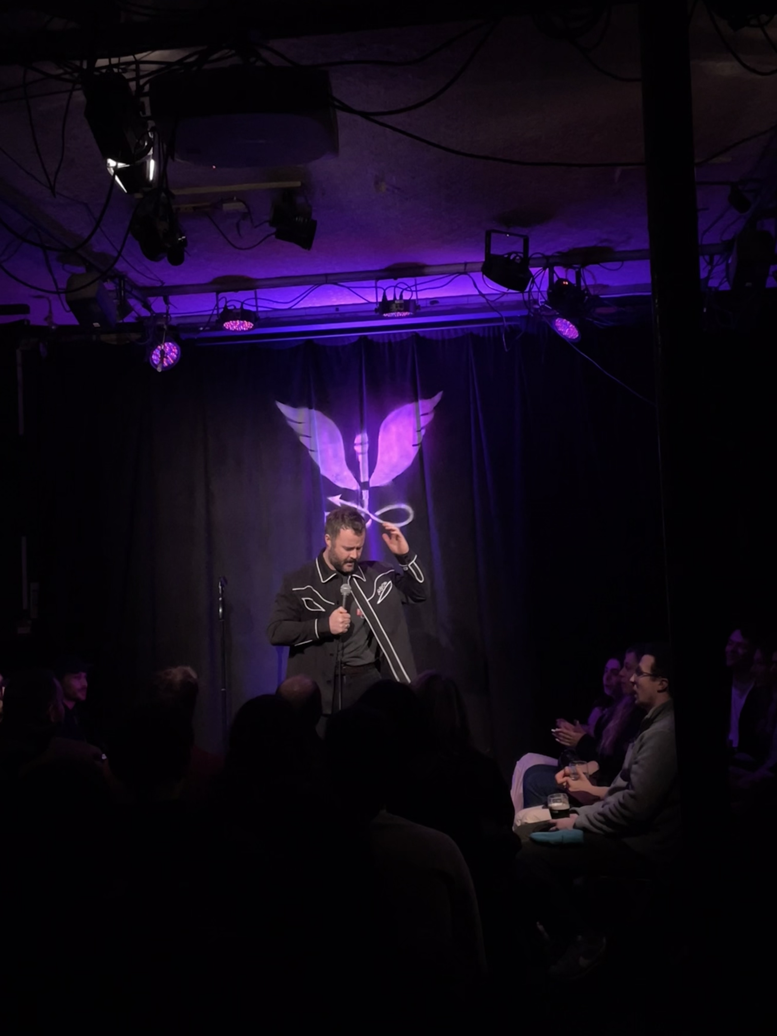A man standing on stage holding a microphone, with a dark curtain and a light projection of a stylized winged symbol behind him, in front of an audience.