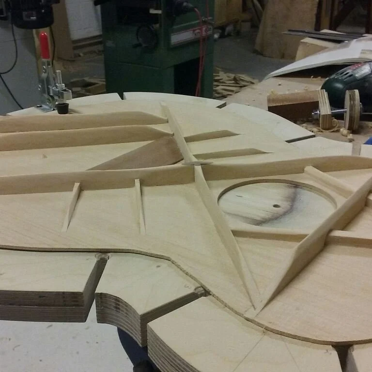 Close-up view of a partially assembled wooden musical instrument, likely an acoustic guitar, in a woodworking workshop.