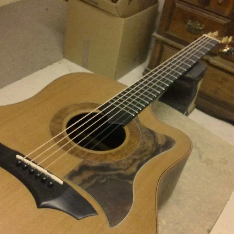 An acoustic guitar with a wooden body and a black pickguard, resting on a surface in a room with furniture and carpet.