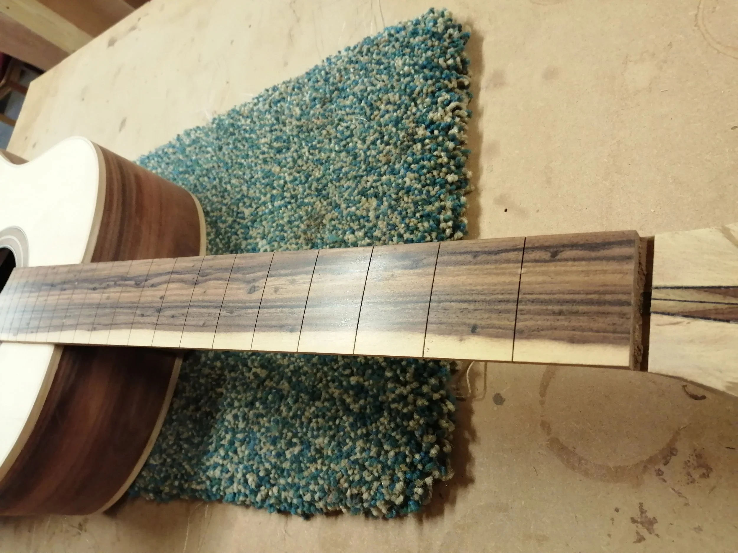 Close-up of a guitar neck with in-progress fret markings, on a workshop table with a textured rug and an acoustic guitar in the background.
