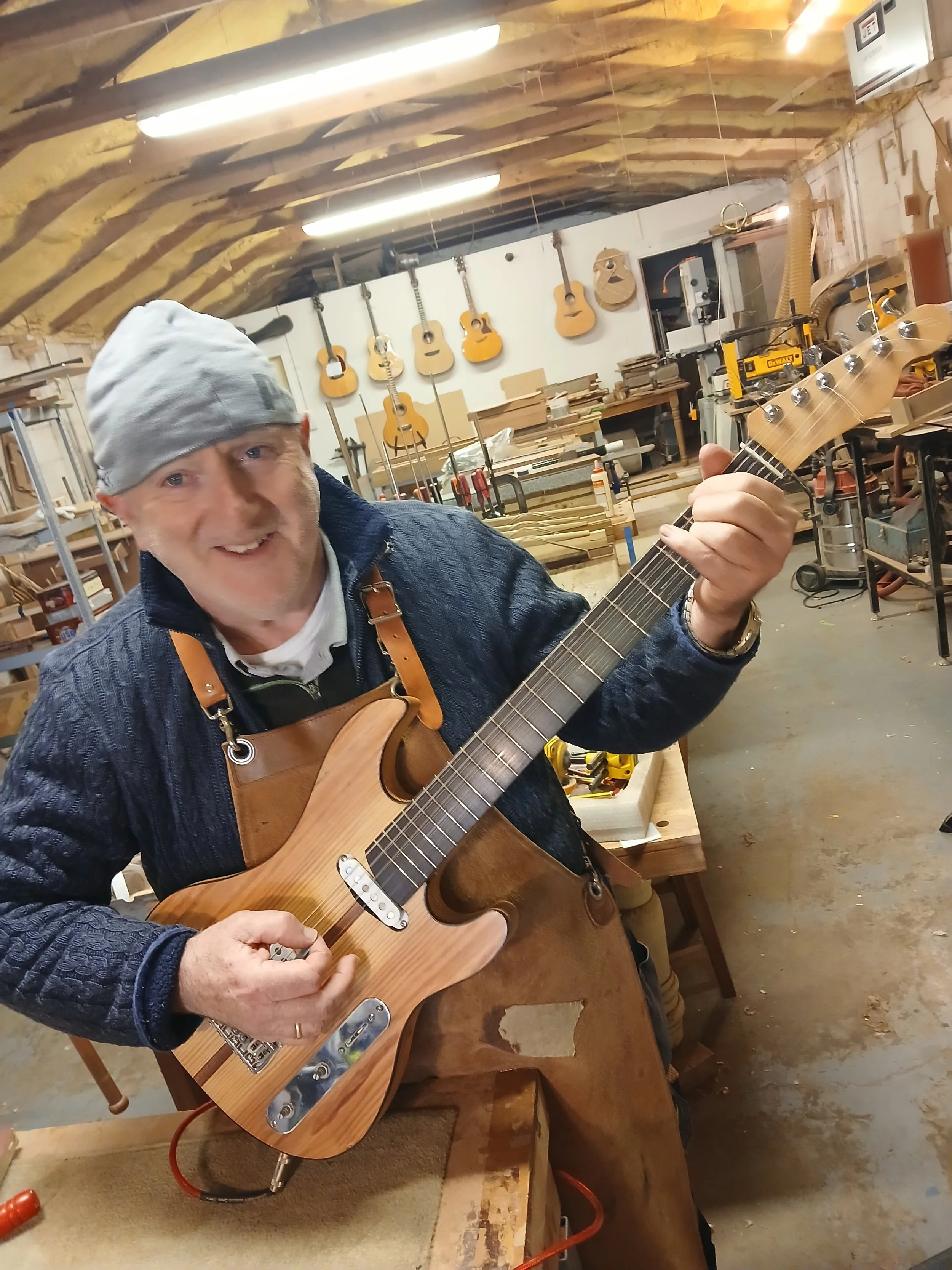 A man in a gray beanie and dark sweater playing an electric guitar in a woodworking shop with guitars hanging on the wall behind him.