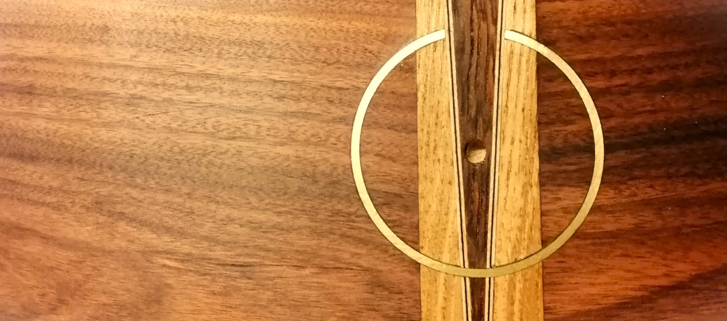 Close-up of a wooden table with a vertical inlay strip and a brass circular metal hinge.