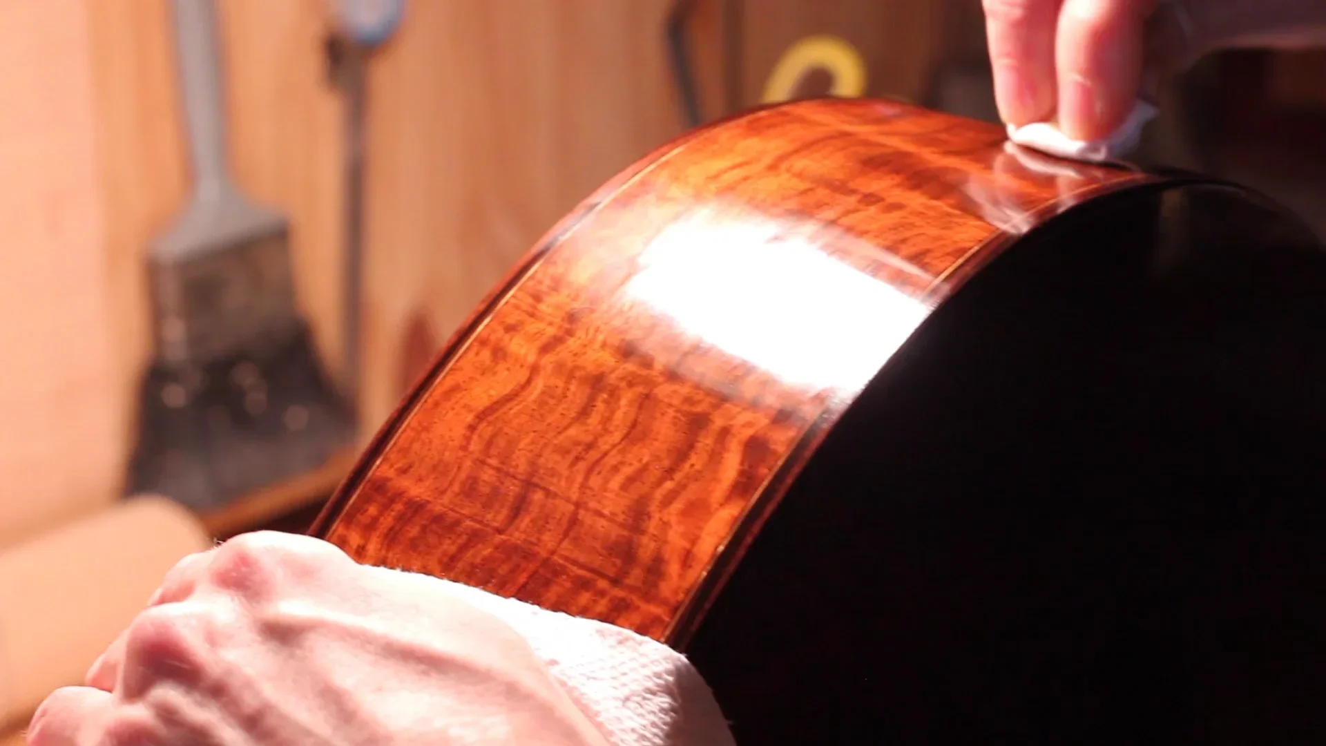 Close-up of a person polishing the back of a wooden acoustic guitar with a white cloth in a workshop setting.