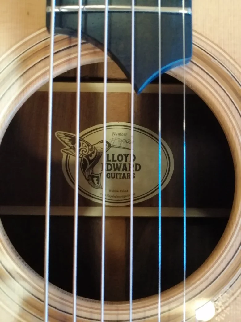 Close-up of an acoustic guitar's soundhole showing the inside label that reads 'Lloyd I. Dwyer Guitars' with a logo of a hummingbird. The strings are visible in the foreground.