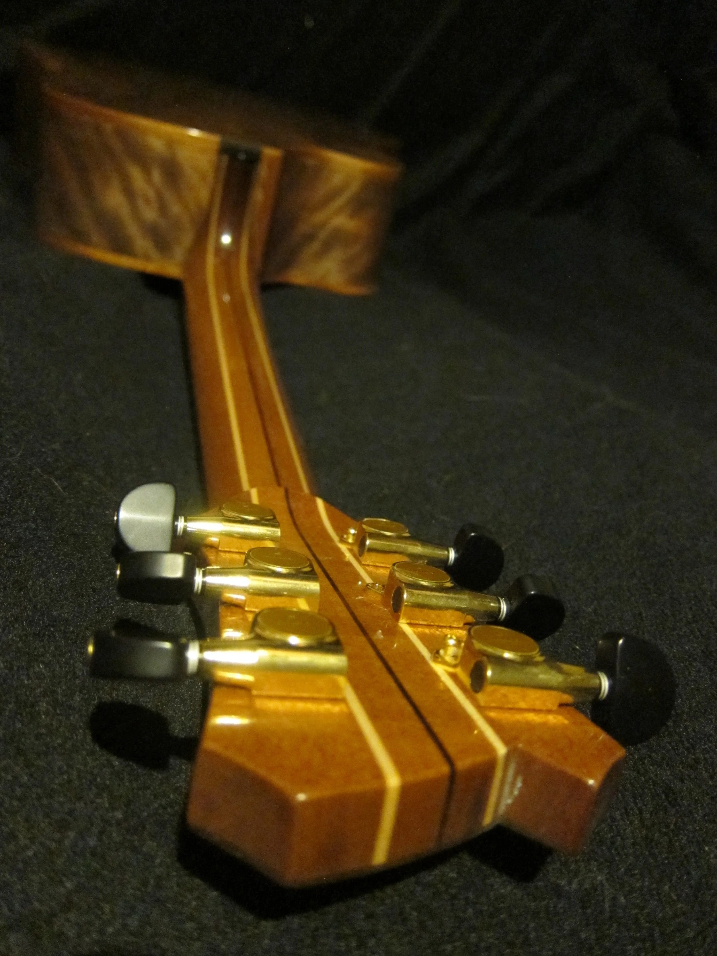 Close-up of the headstock of a wooden guitar or ukulele with tuning pegs, set against a dark background.
