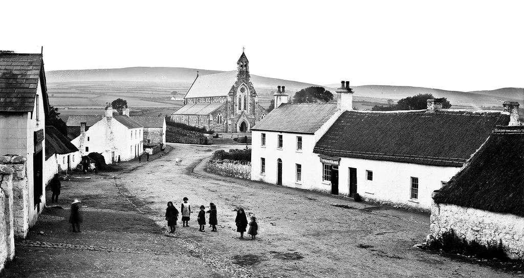 Black and white photo of a village street with old buildings, a church in the background, and children walking.