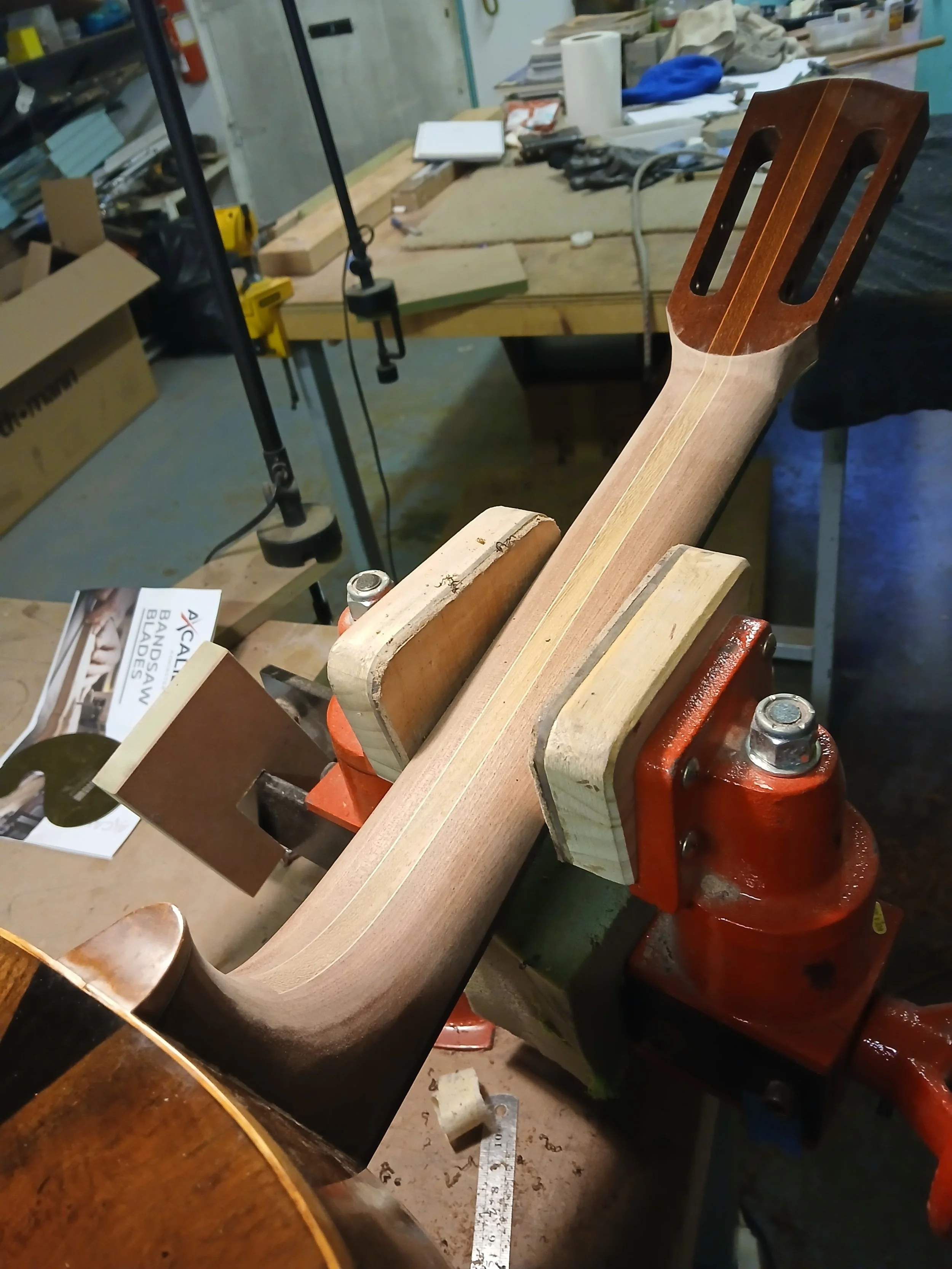 A guitar neck is clamped in a woodworking vice on a workbench in a workshop. The neck is partially sanded and shaped, with the headstock slots cut out.