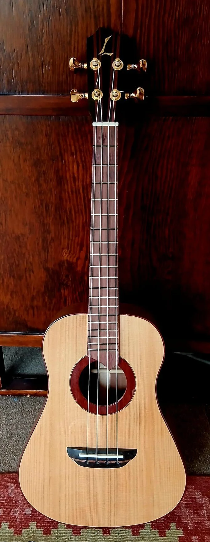 A wooden ukulele with a natural finish, six strings, and gold tuning pegs, resting against a dark wood background.