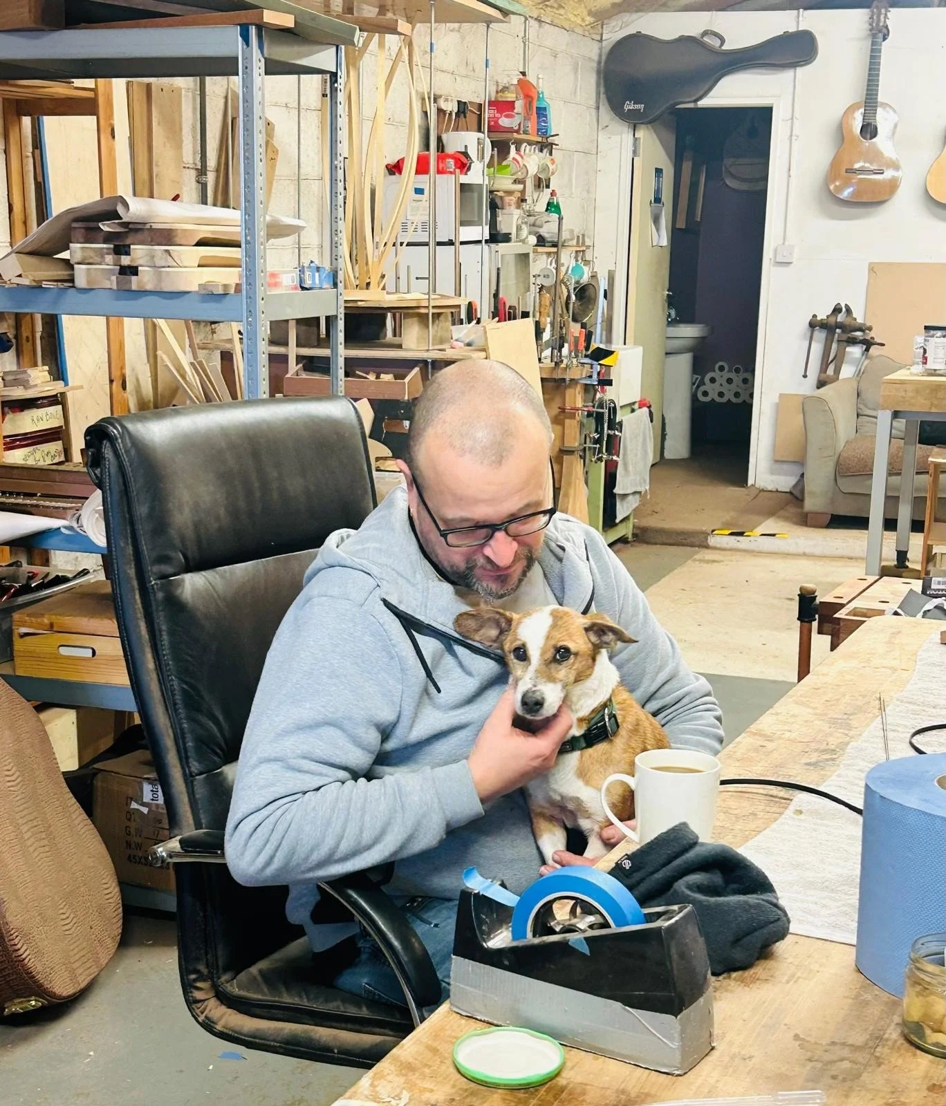 A man sitting at a workbench in a woodworking shop holds a small dog on his lap. The man is wearing glasses and a hoodie, and the dog is looking at the camera. The background shows shelves with woodworking tools and materials.