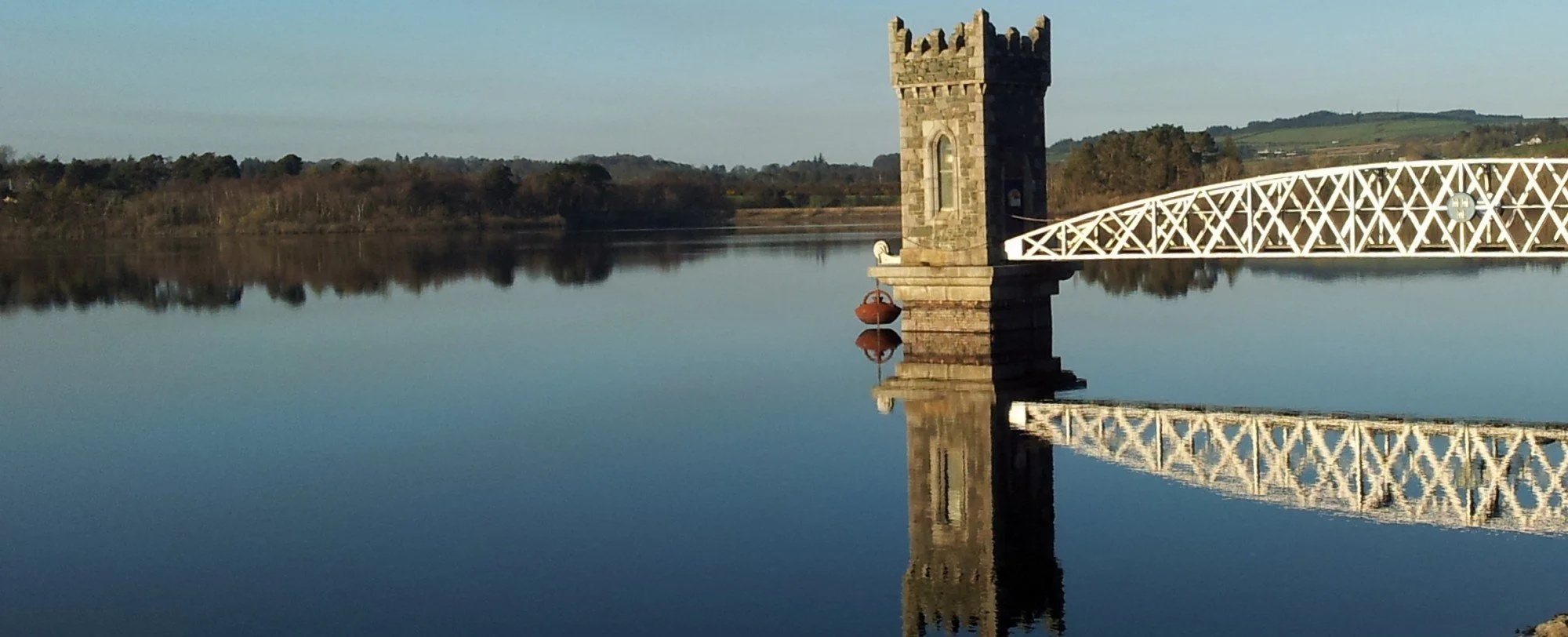 A stone tower and a white lattice bridge over calm water, with reflections visible on the surface, and a background of trees and hills under a clear sky.