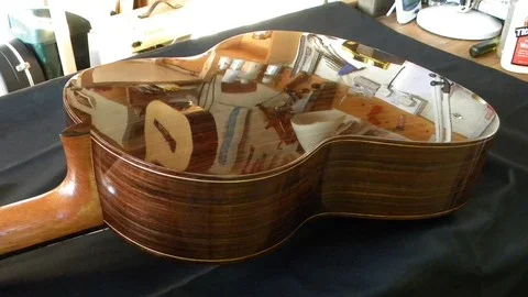 Back side of an acoustic guitar resting on a black cloth with reflections of room items on its polished wooden surface.