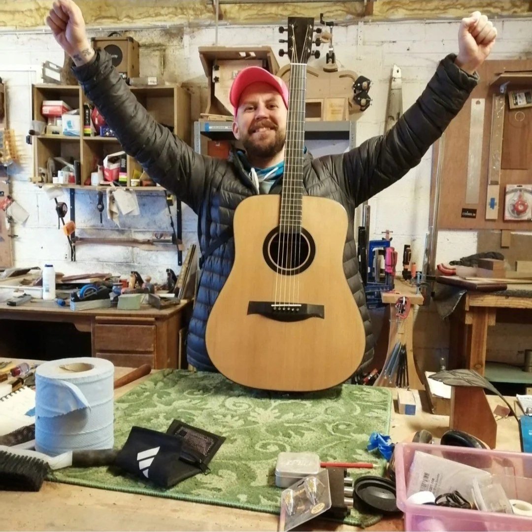 A man wearing a pink cap and black puffer jacket with his arms raised in a woodworking workshop, standing behind an acoustic guitar placed on a green table. The background features shelves with tools, wood pieces, and workshop equipment.