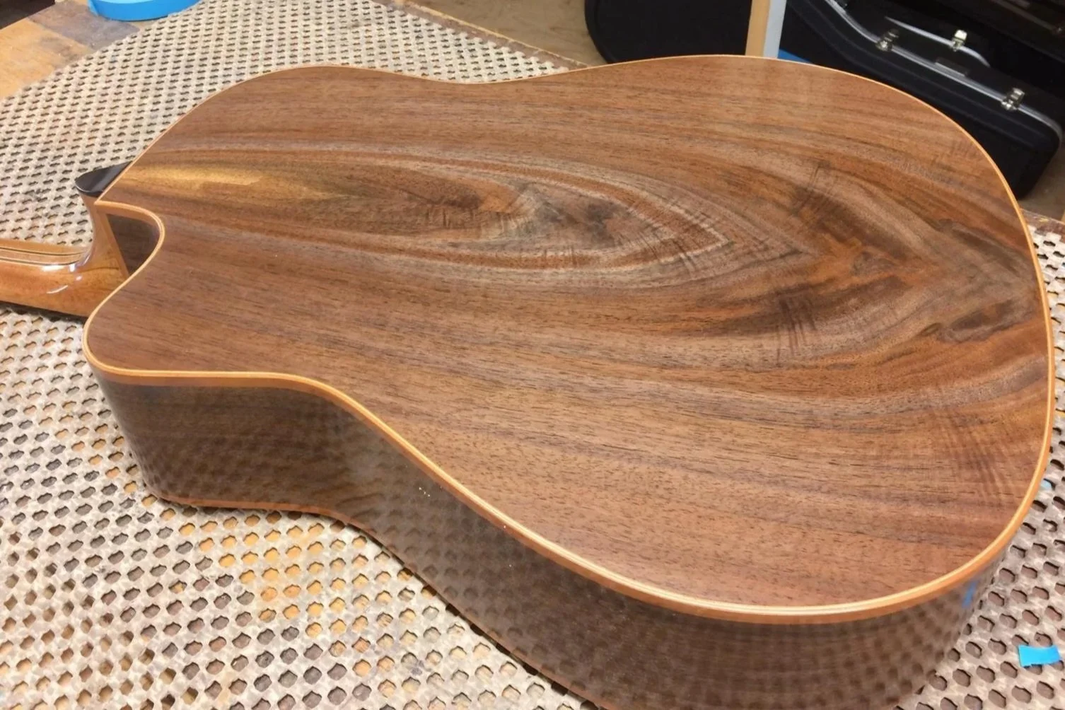 The image shows an acoustic guitar with a wooden body and a natural finish, placed on a textured surface. The guitar's body is seen from the back, highlighting the wood grain and contours.