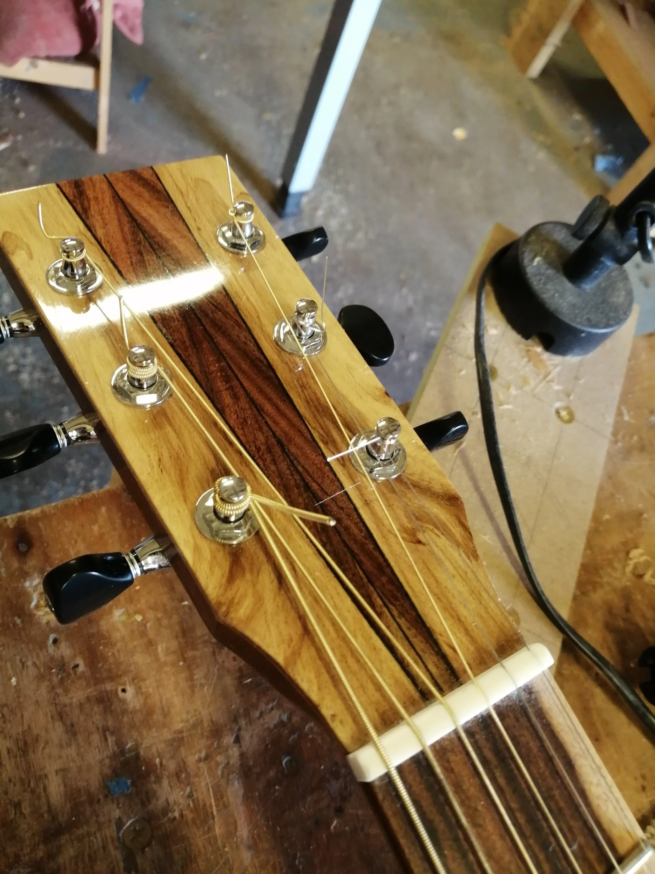 Close-up of a wooden guitar headstock with tuning pegs and strings in a woodworking shop.