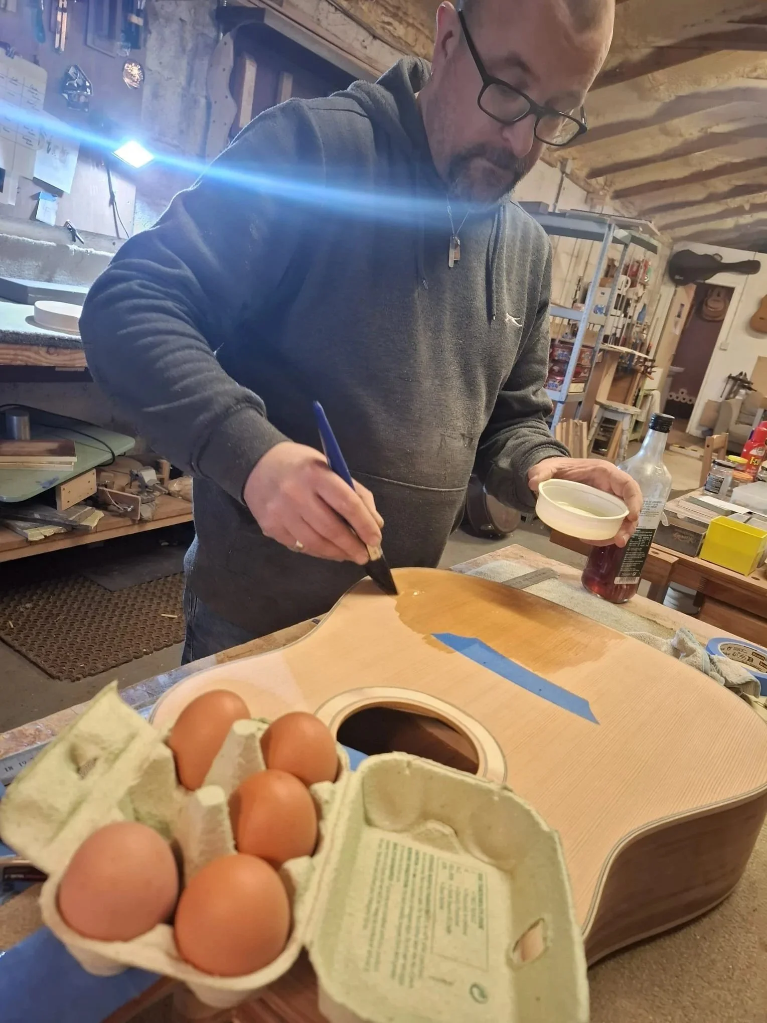 A man with glasses is in a woodworking workshop, painting a guitar body with a brush. Eggs are in an egg carton in the foreground.