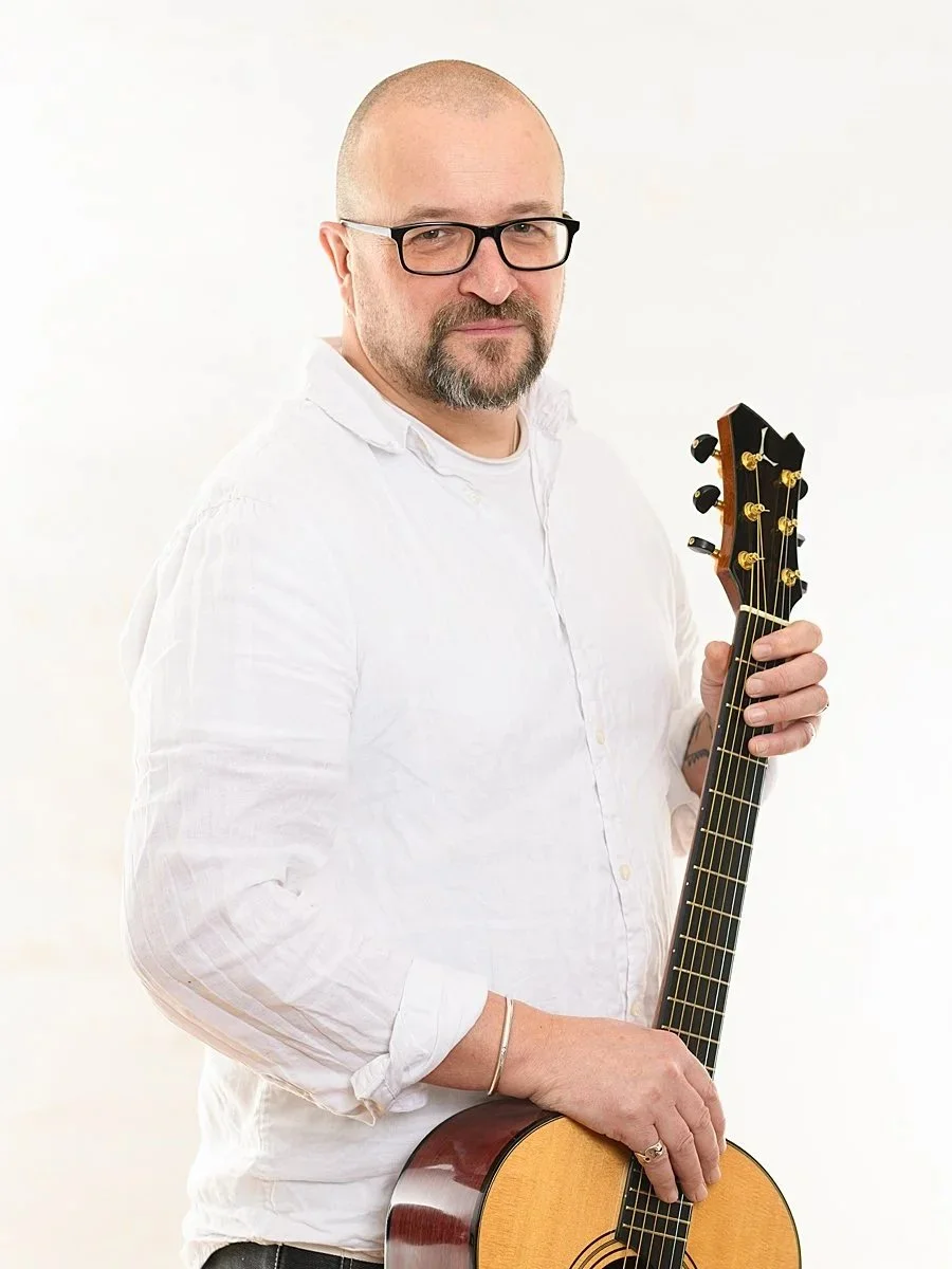 A man with a shaved head, beard, and glasses holding an acoustic guitar against a plain white background.