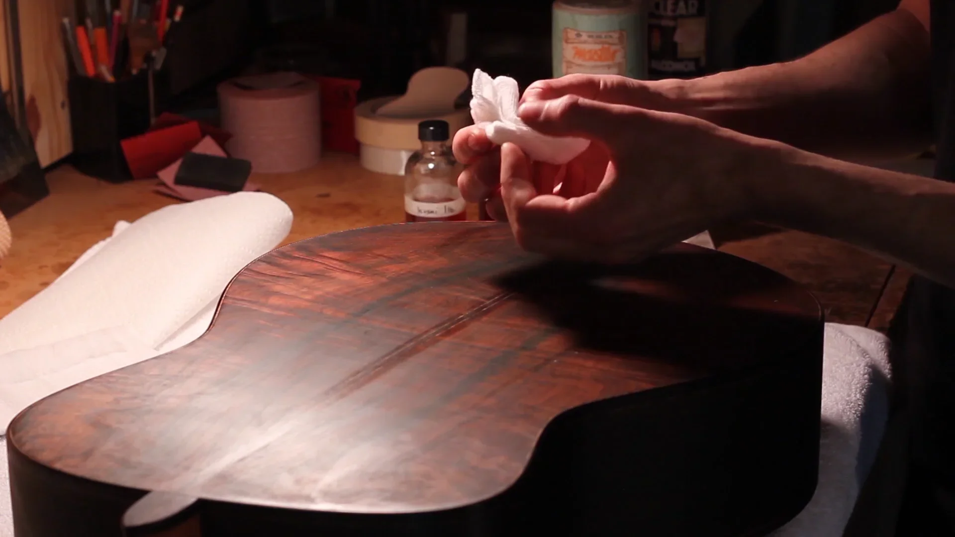 Close-up of a person polishing the back of a wooden acoustic guitar with a cloth on a workbench.