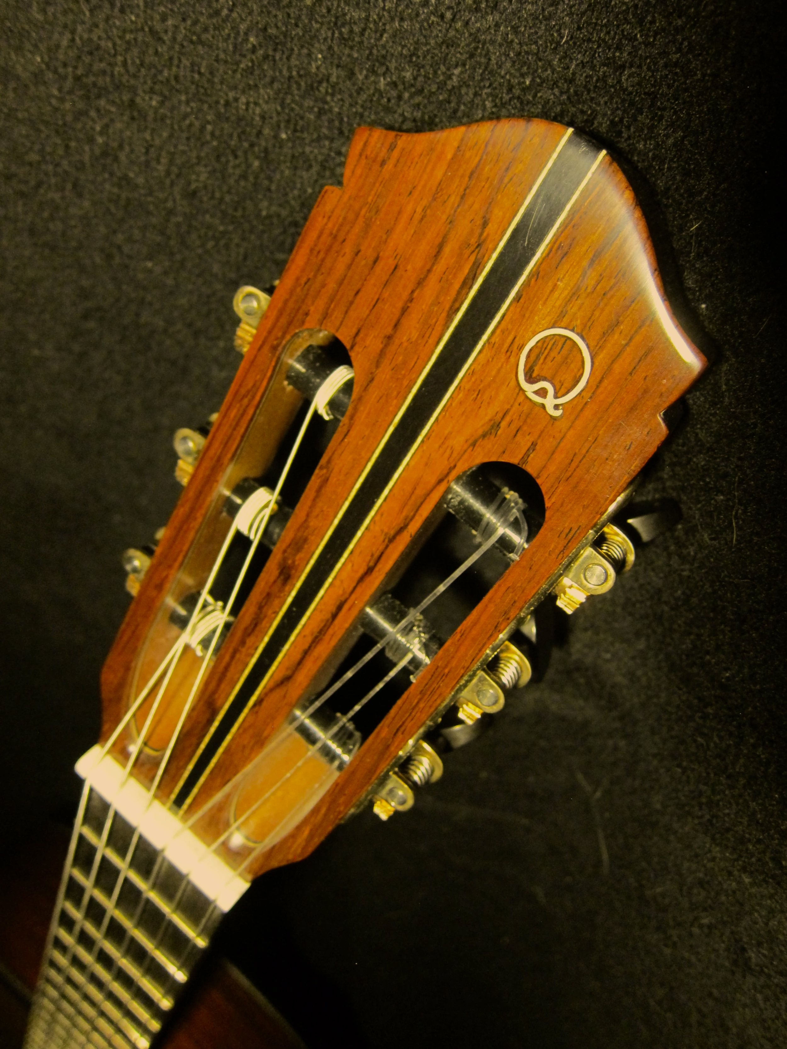 Close-up of a guitar headstock made of wood with tuning pegs and strings, featuring a 'Q' logo.