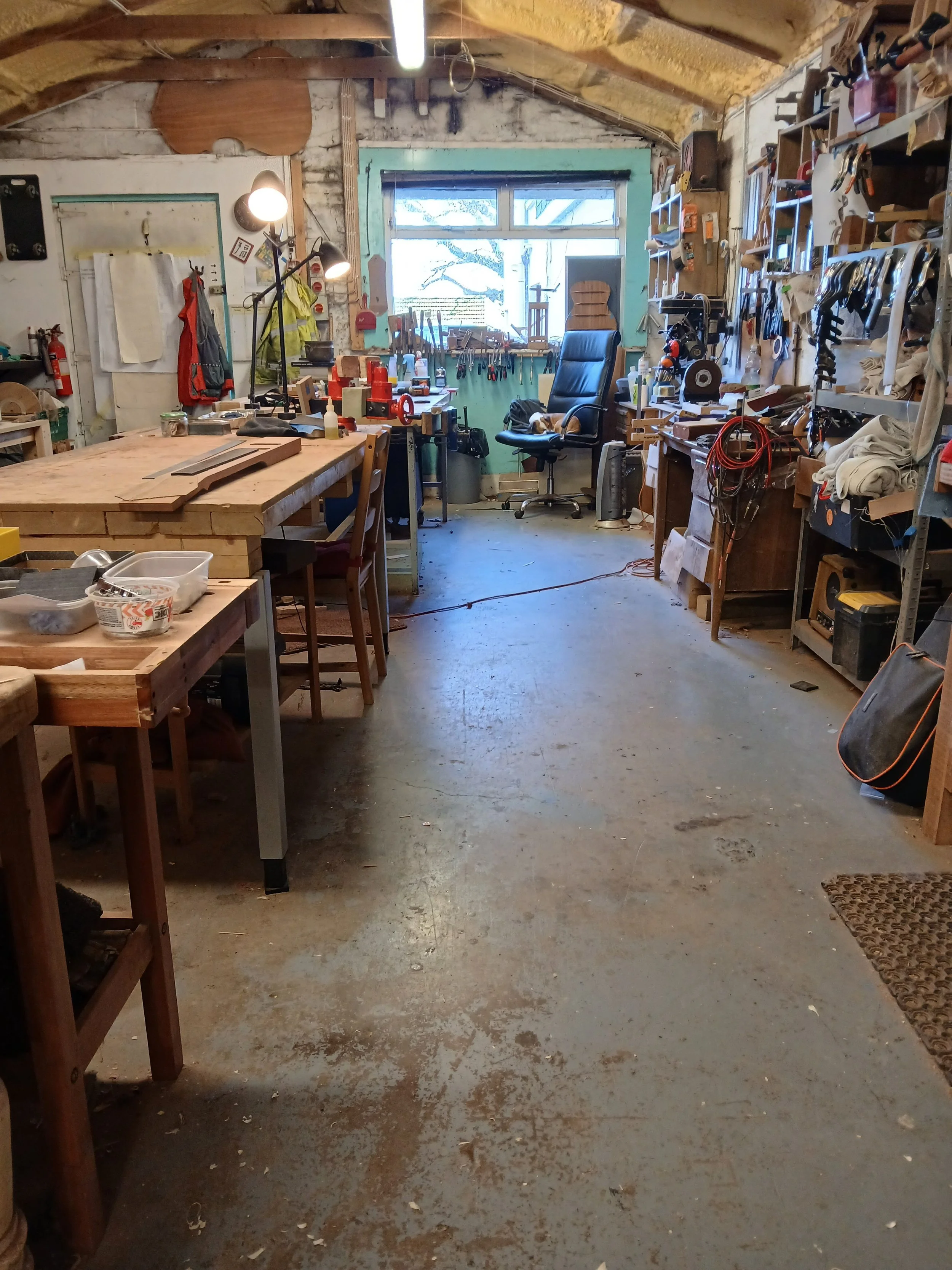 A woodworking workshop with workbenches, tools, shelves, and a window at the back letting in natural light.