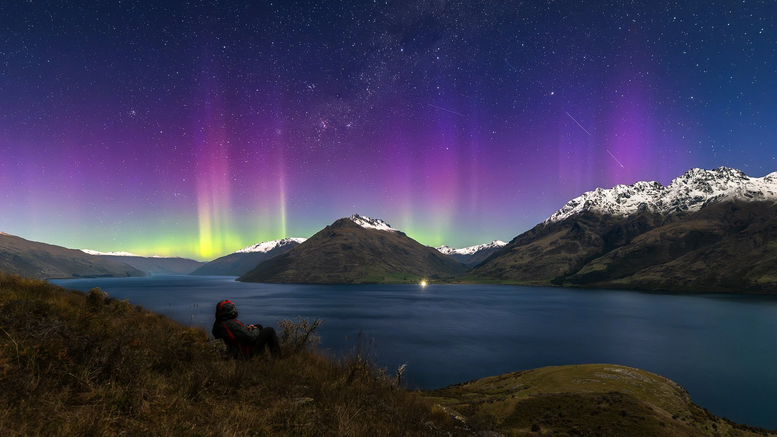 Aurora Australis glowing in vibrant purple, pink, and green over Lake Wakatipu and snow-capped Southern Alps, New Zealand, with a photographer looking out.