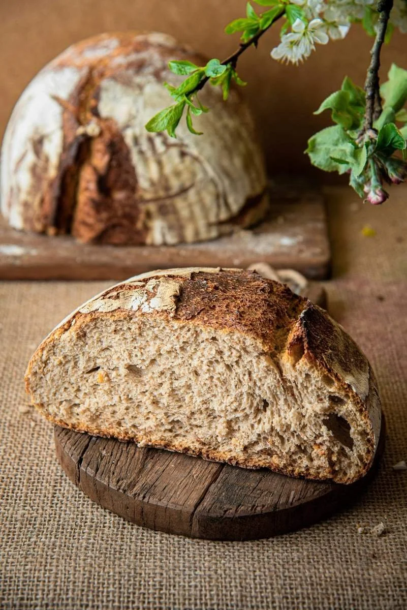 A loaf of rustic bread with a crusty top, partially sliced, resting on a round wooden cutting board. In the background, there is another loaf of bread and a branch with green leaves and white blossoms, all set on a textured surface.