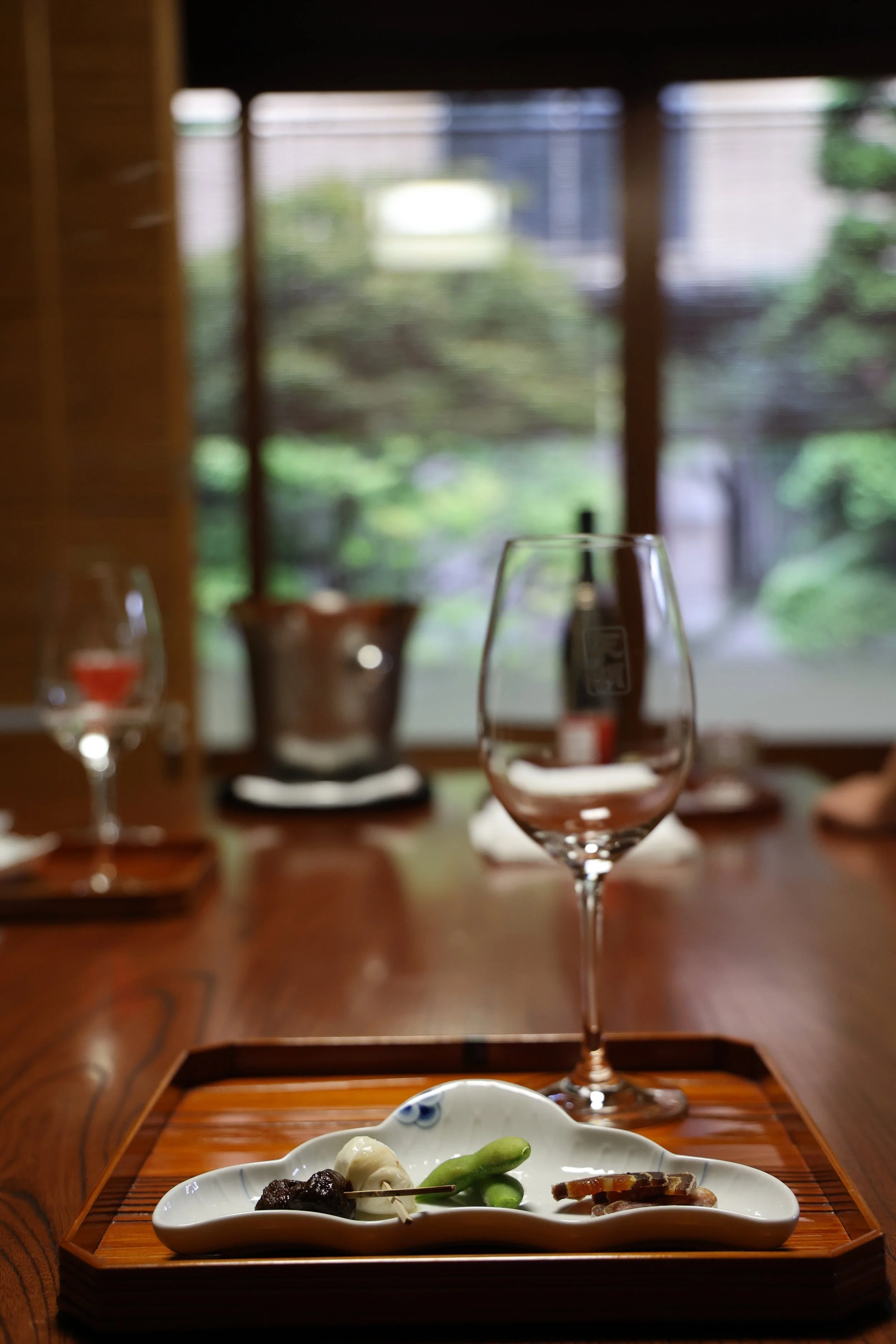 Empty wine glass in focus on a wooden table with a small white dish of assorted appetizers, including green beans, a skewer, and a dark paste, in a restaurant with a window showing trees outside.