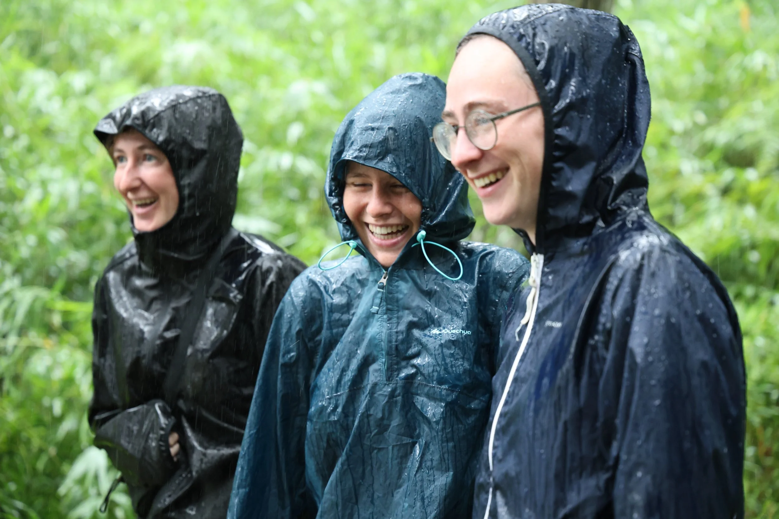 Three people wearing rain jackets and hoods, smiling and laughing in a green forest during rainy weather.