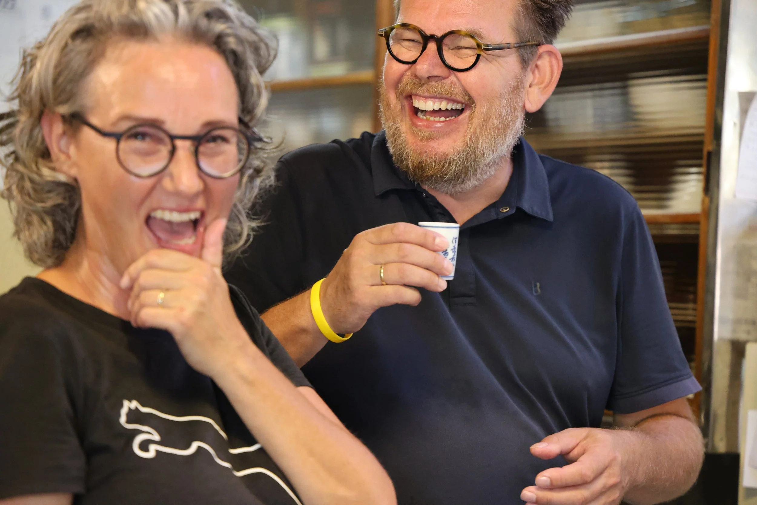 Two people laughing together, a woman with curly gray hair and glasses, and a man with glasses and a beard, holding a small cup.