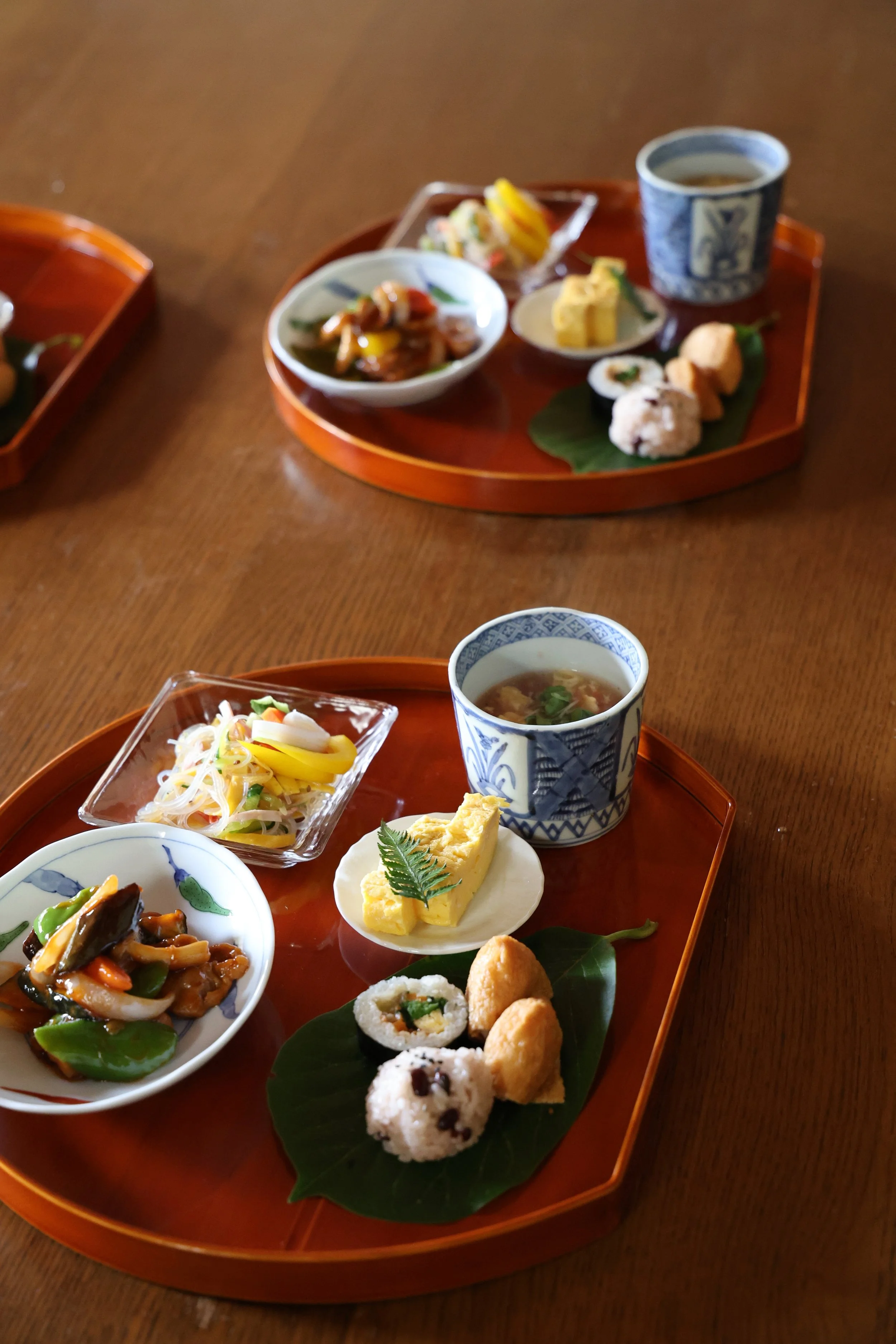 Two trays of traditional Japanese cuisine with rice balls, pickled vegetables, soup, tamagoyaki, and sushi, arranged on a wooden table.