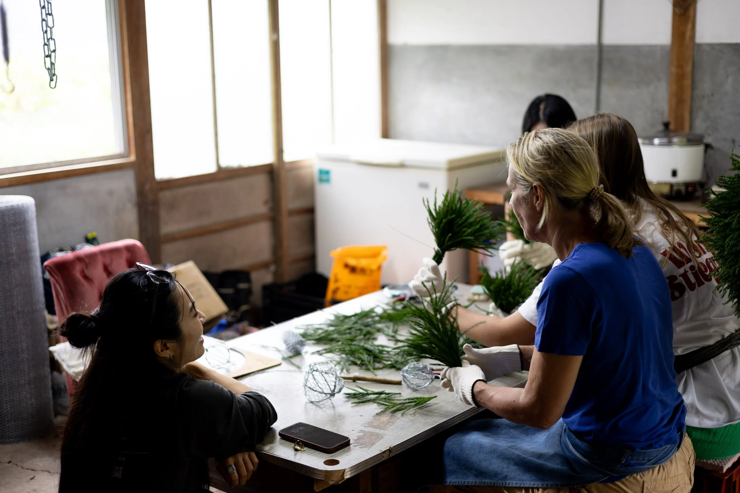 People making Christmas centerpieces with pine branches at a workshop table in a rustic room.