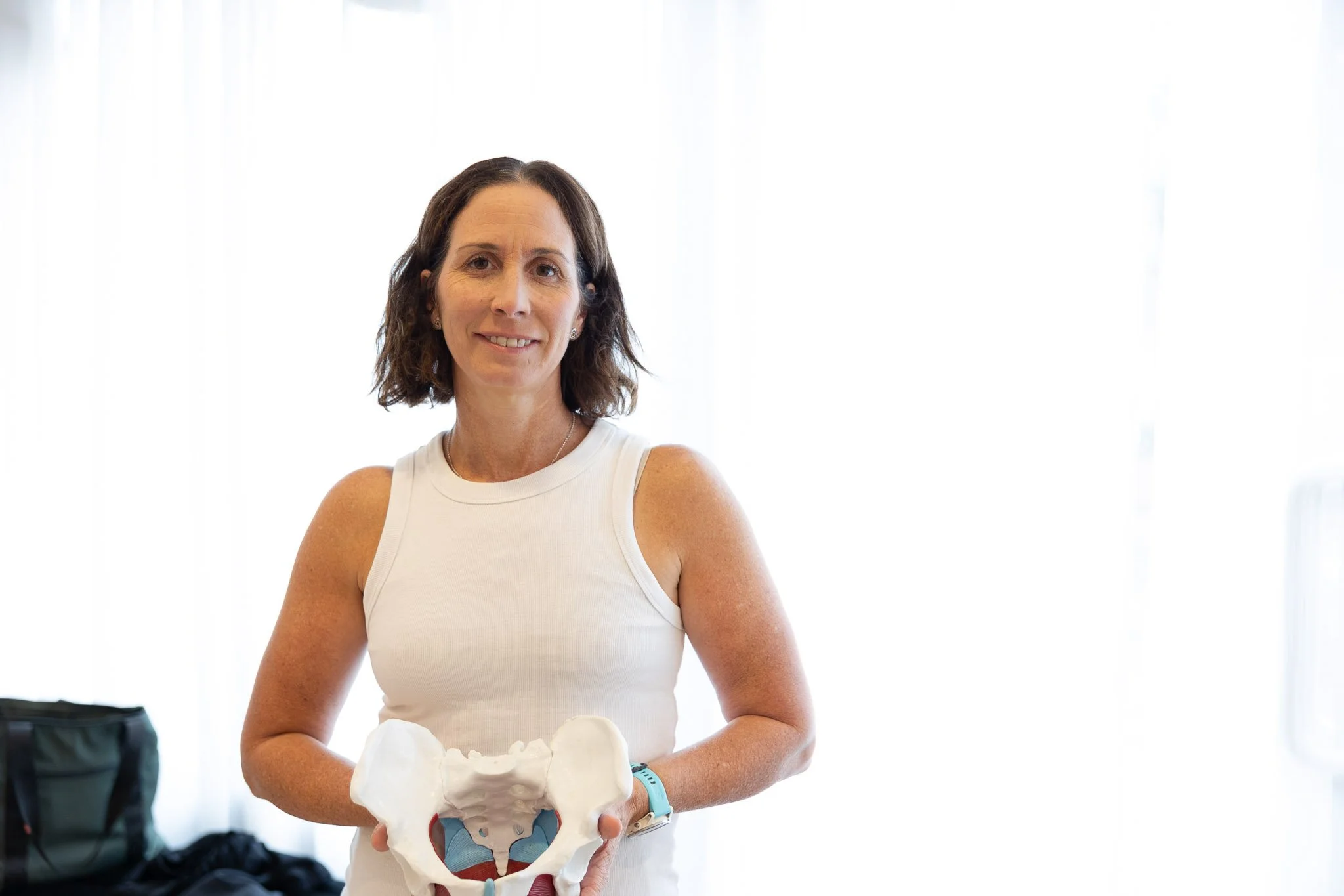 A woman with shoulder-length brown hair, wearing a white sleeveless top, holds a model of a human pelvis in front of her. She is standing indoors near a window with bright natural light, with a black bag and other items visible in the background.