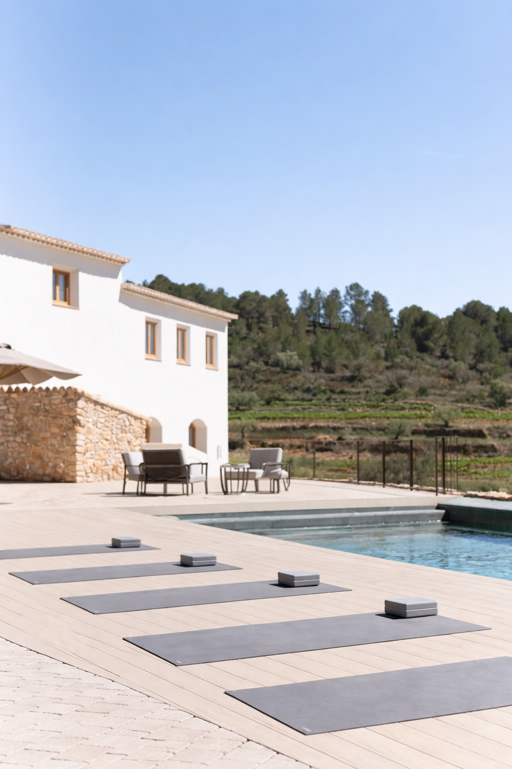 Setting up for outdoor yoga class with six yoga mats and blocks near a swimming pool, with a white house and hilly landscape in the background under a clear blue sky.