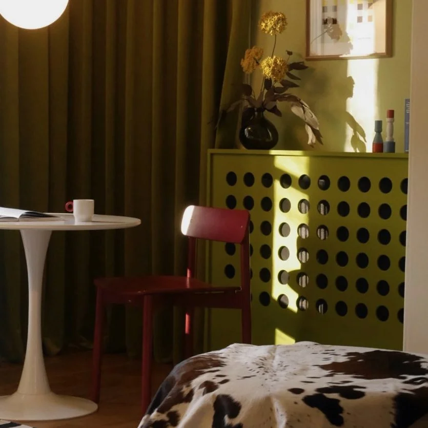 Interior scene with a white round table, a red chair, a yellow radiator cover with circular cutouts, a black vase with yellow flowers, and a cowhide blanket in the foreground. Sunlight creates shadows on the wall.