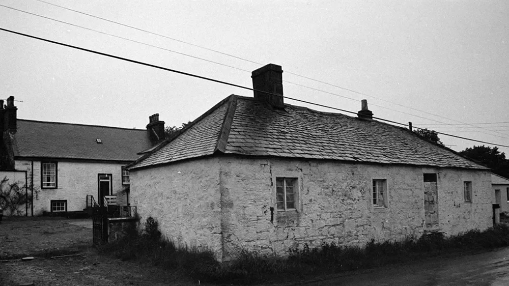 Older white stone house with a sloped roof and chimney, two small windows, situated next to a modern house, on a street with utility wires overhead in a rural setting.