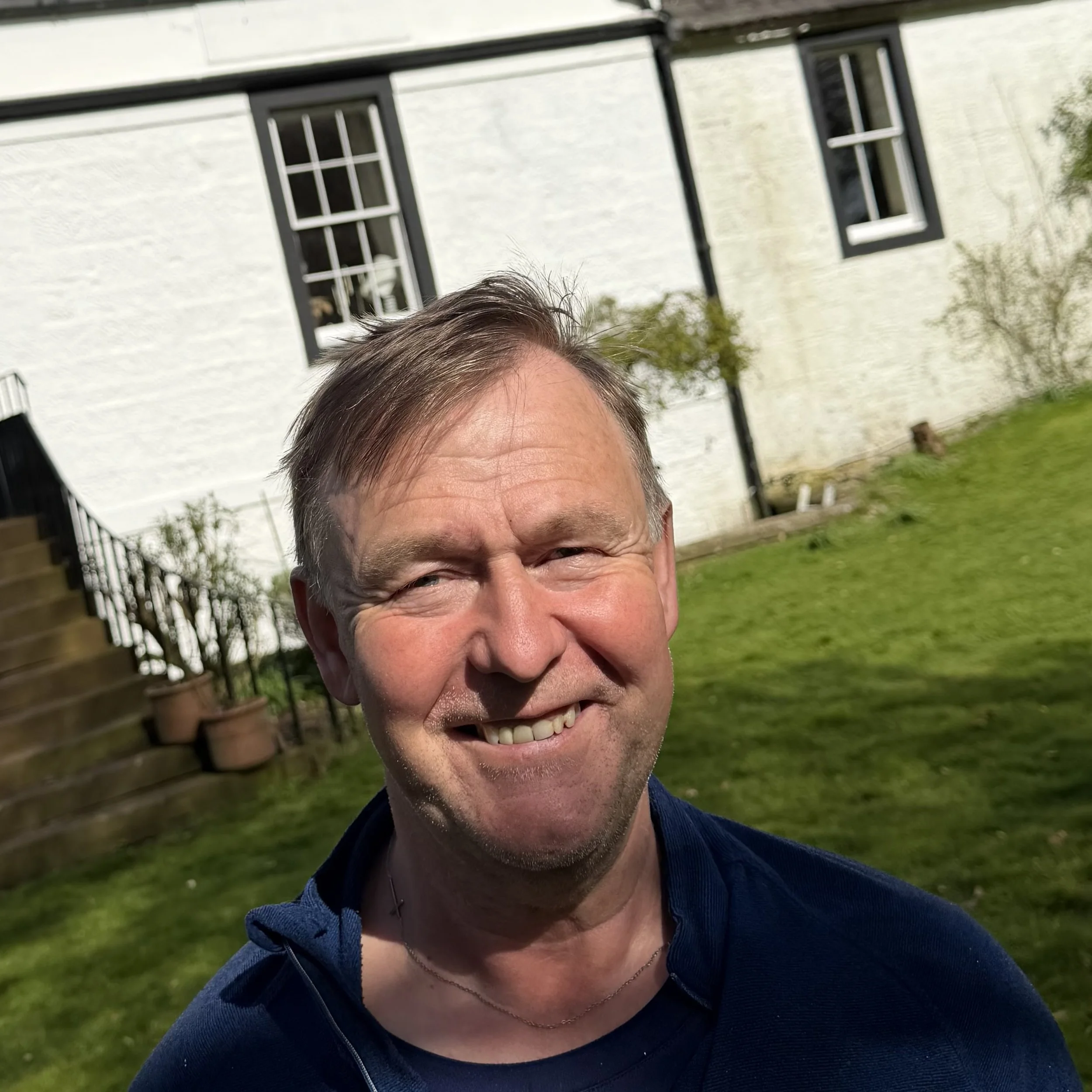 Close-up of a man smiling outdoors, with a house and garden in the background.