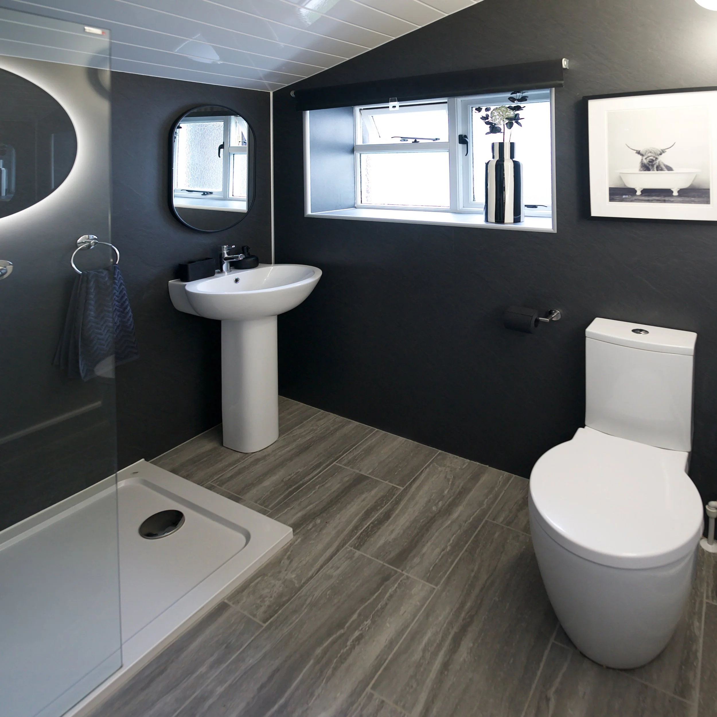 Modern bathroom with dark gray textured walls, a white pedestal sink with a black soap dispenser, circular mirrors on the wall, and a window with a sill holding a decorative vase with flowers. A framed black-and-white photograph of a cow in a bathtub decorates the wall. The floor is covered with gray wood-look tiles, and a white shower tray is visible at the bottom of the image.