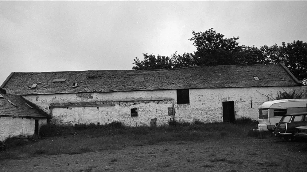 Older white brick building with a pitched roof, some windows, and a door, surrounded by grass and some trees in the background. Part of a vintage camper trailer and a car are visible on the right side.