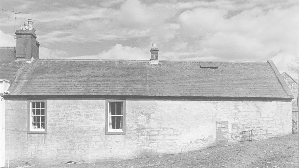 A black and white photo of an old stone house with a sloped roof, two visible windows, and a small door, under a partly cloudy sky.