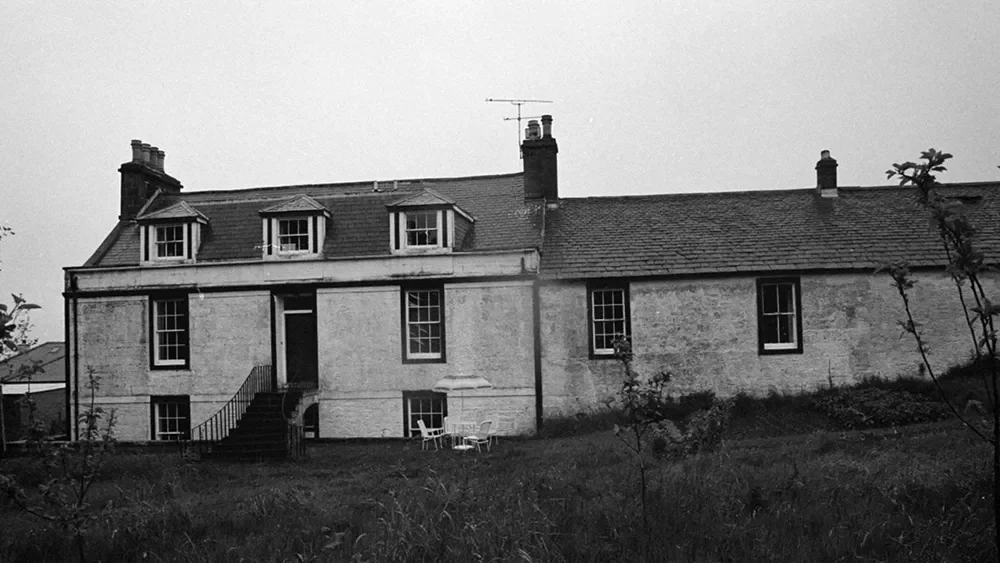 Black and white photo of a large, old house with multiple chimneys and small dormer windows on the roof, surrounded by a grassy area with some plants and trees.