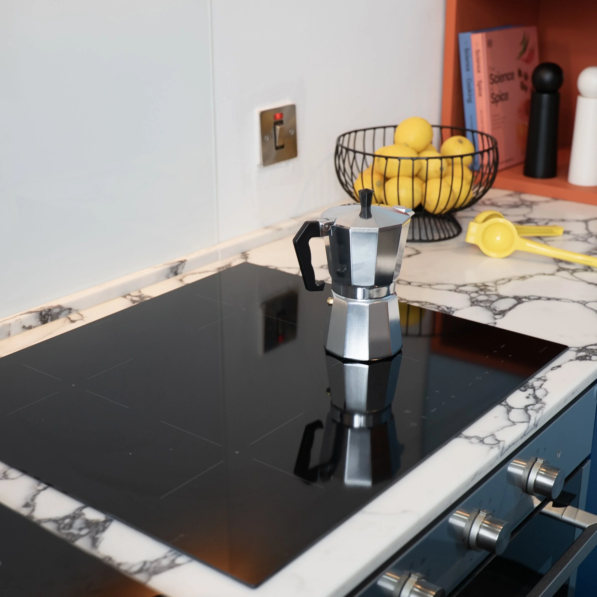 Close-up of a coffee maker on a marble countertop in a kitchen, with a wire basket of lemons, cookbooks, and black and white pepper shakers in the background.